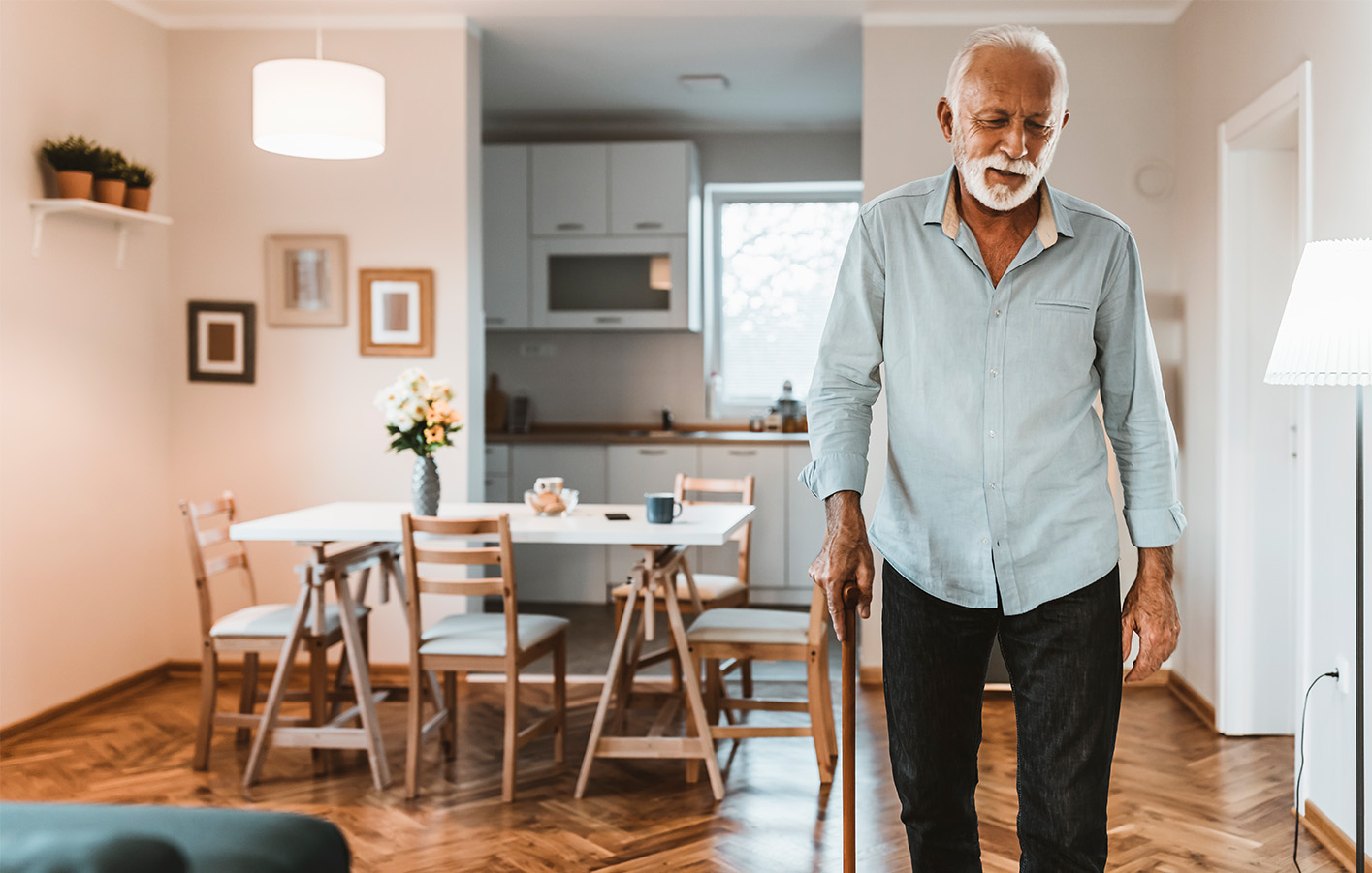 Senior geht mit Krückstock durch das Wohnzimmer Älterer Herr mit weißem Haar und Krückstock geht vorsichtig durch ein helles Wohnzimmer mit Essbereich.